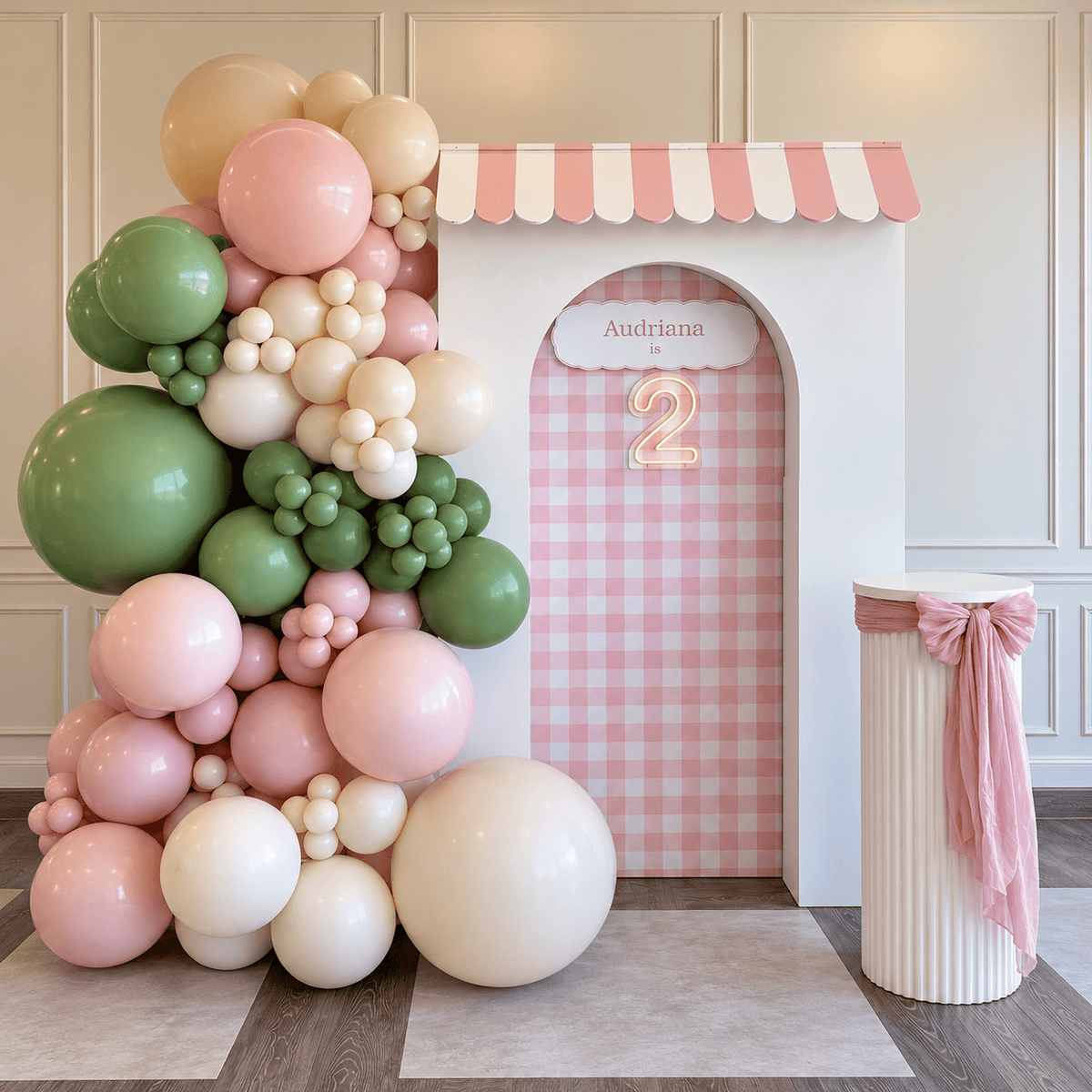 Pink gingham second birthday backdrop with custom storefront awning, pastel pink sage green and ivory balloon garland, illuminated number 2 and white cake pedestal with pink bow.