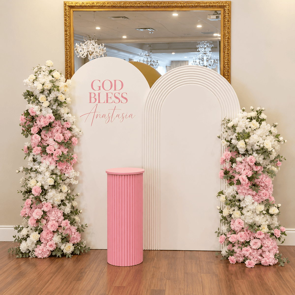 Pink and white baptism backdrop with two white arch panels, personalized God Bless lettering, two large artificial floral arrangements and pink ribbed cake pedestal.
