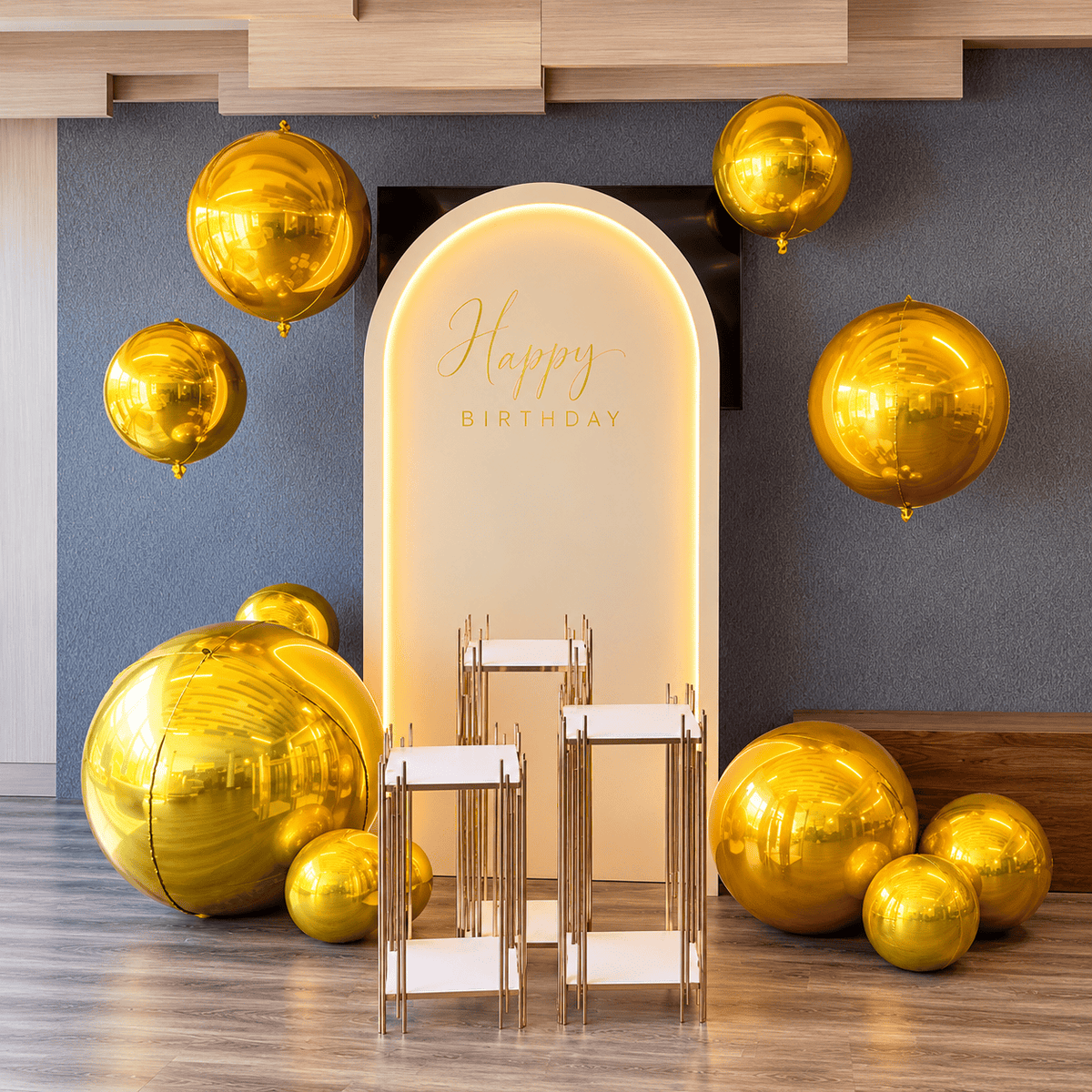 Luxury gold chrome birthday balloon decor with illuminated arch backdrop, oversized mirror gold balloons, custom Happy Birthday lettering and three gold display pedestals.