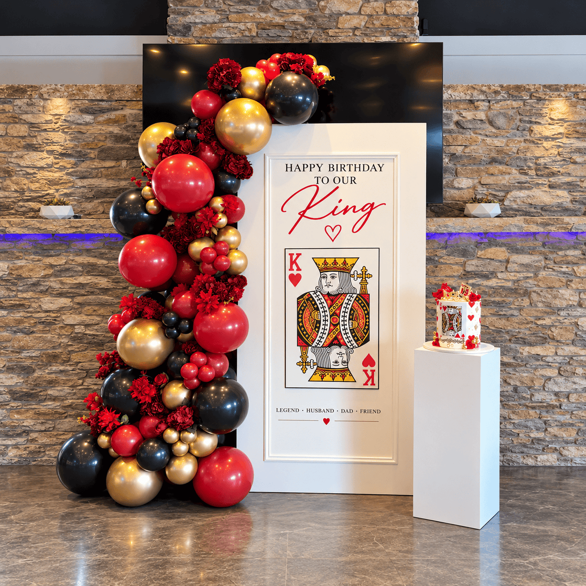 King of Hearts birthday backdrop with custom playing card design, red black and gold balloon garland, red flowers and white cake pedestal.