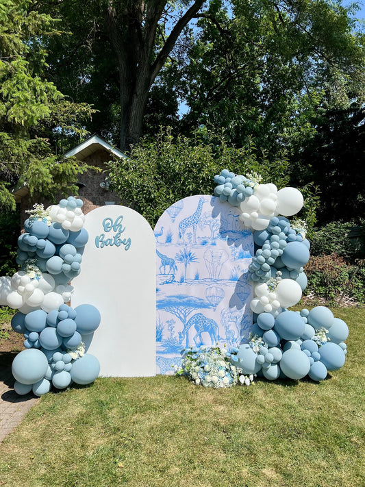 Blue Safari baby shower backdrop with white and safari-printed walls, blue & white balloon garlands, Oh Baby sign, and floral décor
