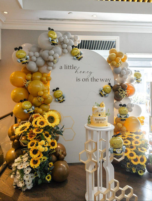 Bee theme baby shower backdrop with white oval wall, sunflowers, daisies, bee figures, honeycomb décor, balloon garlands, and BABEE boxes