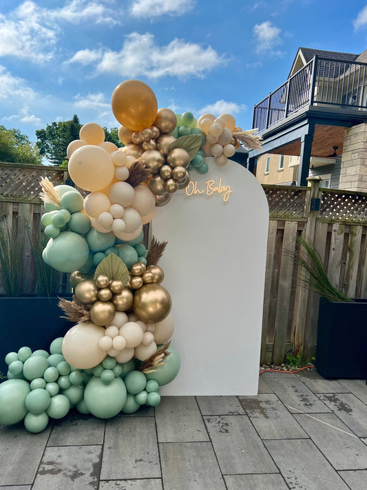 White oval baby shower backdrop with Oh Baby neon sign, beige gold sage green balloon garland, and dried florals