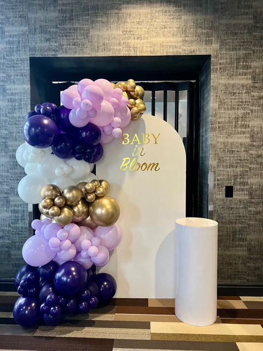 Baby in Bloom baby shower backdrop with gold inscription, purple lavender white & gold balloon garland, and cake table