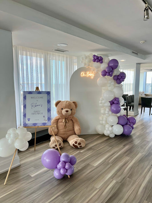 Teddy bear baby shower backdrop with white oval wall, white and lilac balloon garland, Oh Baby neon sign, teddy bear décor, and welcome sign