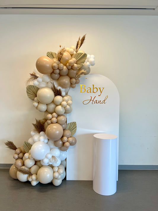 Boho baby shower backdrop with white oval wall, neutral balloon garland, dried florals, palm leaves & cake table