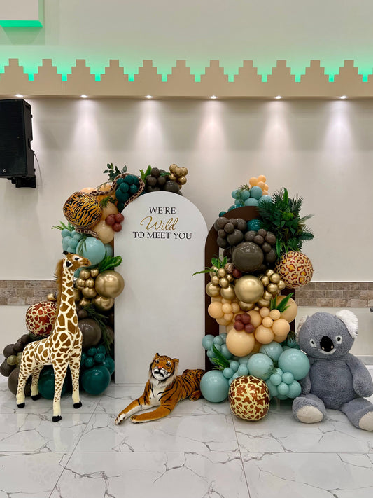 Safari baby shower backdrop with white and brown arches, balloon garlands, greenery, giraffe, tiger, snake, koala & custom inscription