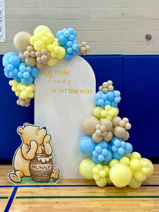 Winnie the Pooh baby shower backdrop with white oval wall, balloon garlands, and Pooh cut-out with honey pot décor