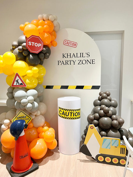 Construction theme birthday backdrop with white oval backdrop, balloon garlands, cake table, excavator cut-out, cones, and hard hat props