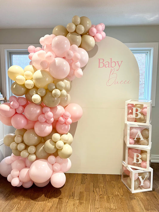 Baby Shower setup with white oval backdrop, pastel pink and beige balloon garland, and Baby Boxes