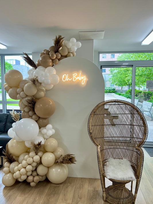 Neutral baby shower backdrop with white oval wall, beige balloon garland with dried florals, Oh Baby neon sign & peacock chair
