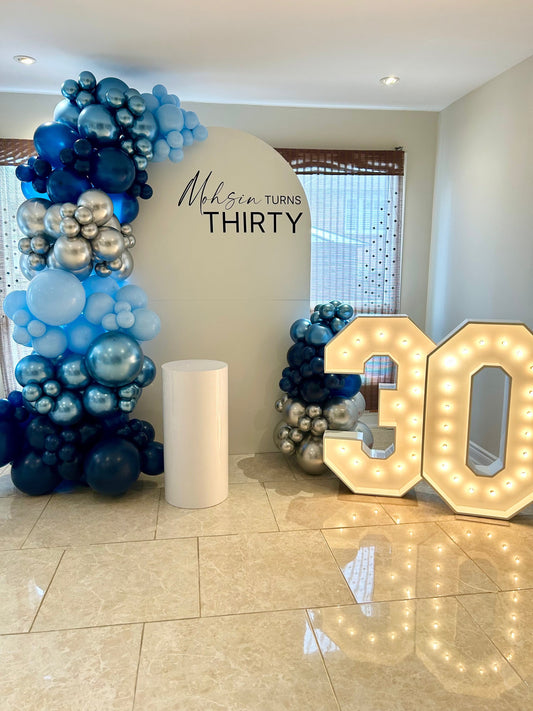 White oval birthday backdrop with custom inscription, blue & silver balloon garlands, marquee numbers 30, and white cake table