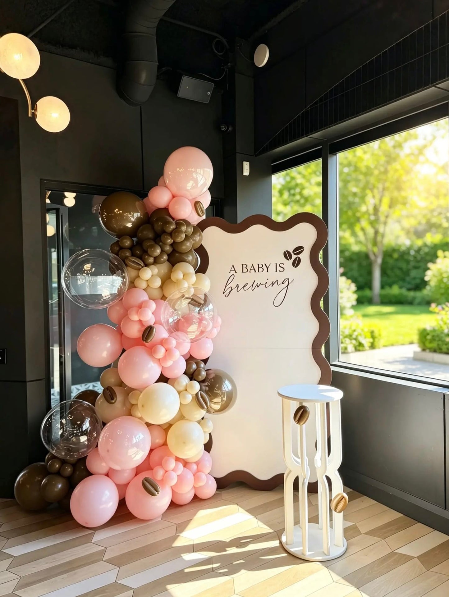 A Baby Is Brewing coffee theme baby shower backdrop with pink, cream and brown balloon garland, clear bubble balloons, coffee bean accents and white cake table.