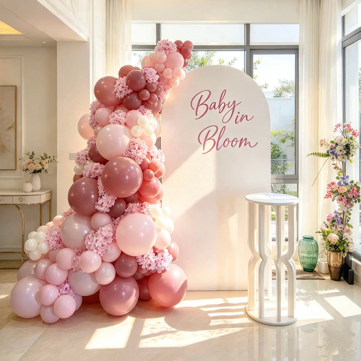 Baby in Bloom baby shower backdrop with white arch, pink and mauve balloon garland, pink floral accents, personalized lettering and white cake pedestal.