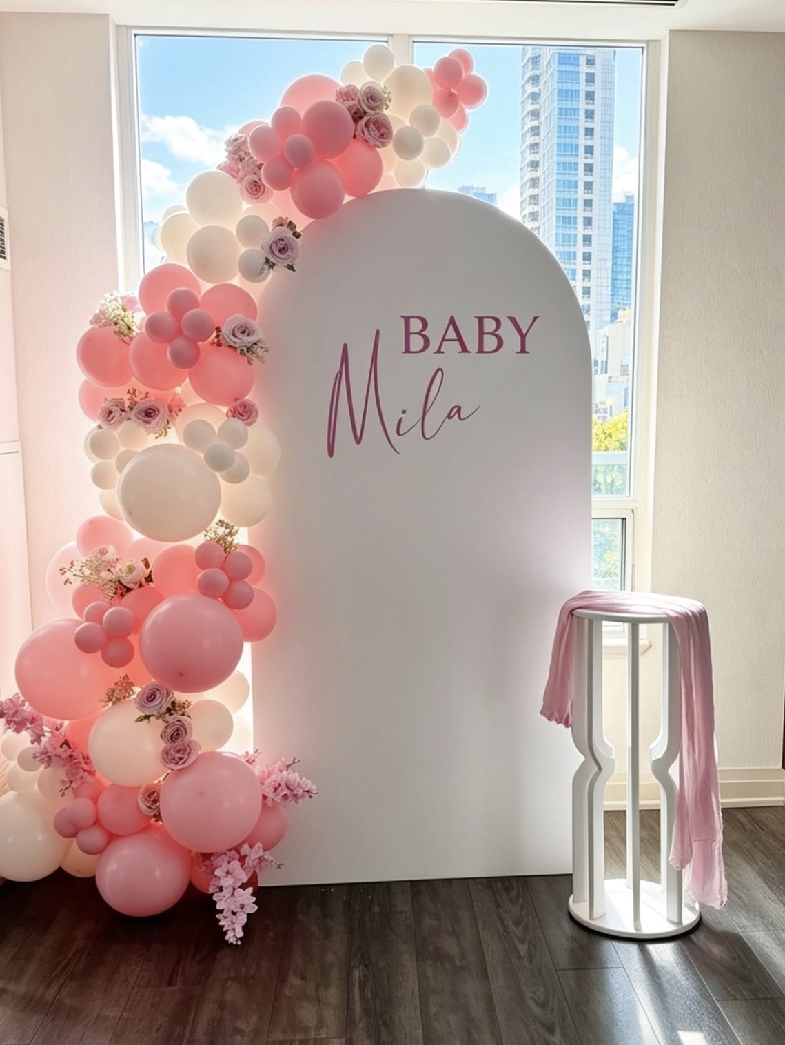 White arch backdrop with blush pink and ivory balloon garland, floral accents and custom baby girl signage.