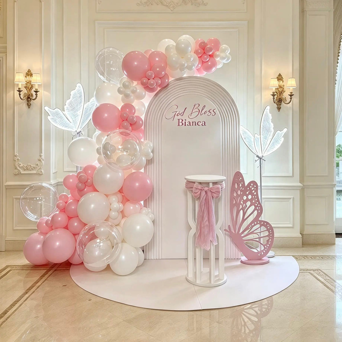 Pink and white baptism backdrop with ripple arch, personalized God Bless lettering, organic balloon garland, transparent bubble balloons, illuminated butterflies, pink butterfly cutout and cake pedestal.