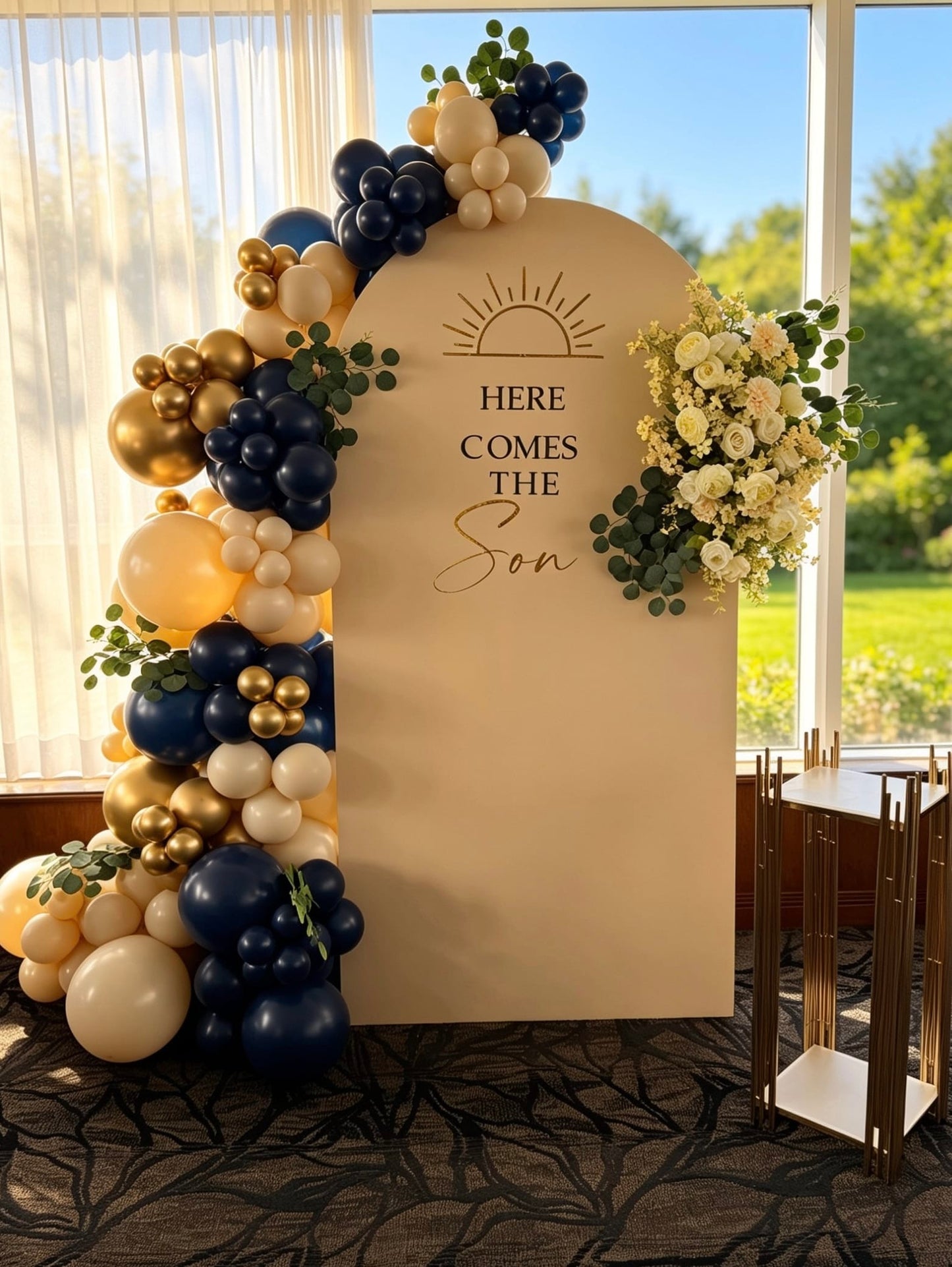 Beige baby shower backdrop with a navy blue, ivory and gold balloon garland, greenery, coordinating floral arrangement and a modern gold display stand.