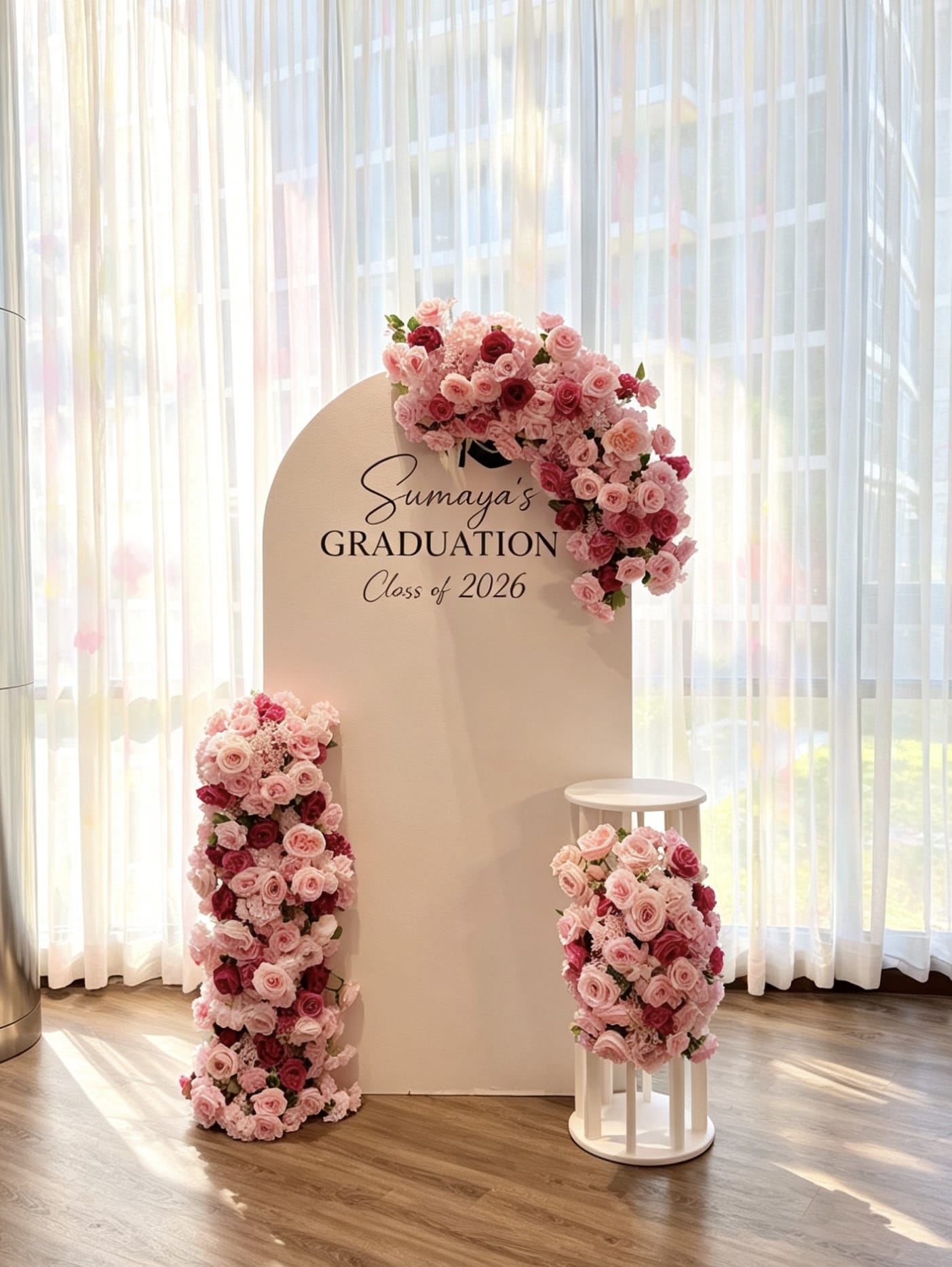 Graduation backdrop featuring a white arch panel with custom lettering, luxurious pink faux floral arrangements and a white pedestal stand.