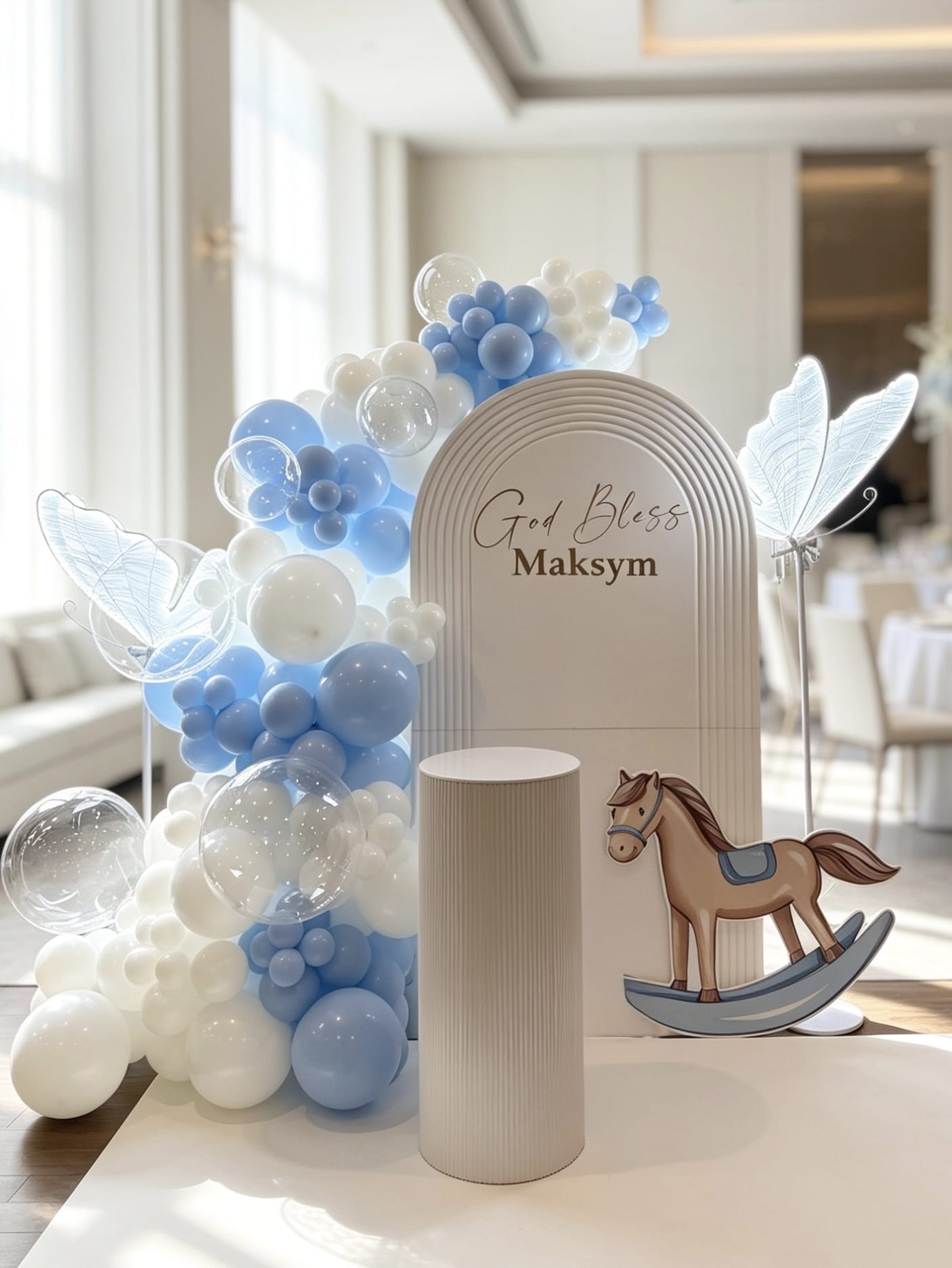Blue and white baptism backdrop with a ripple arch, bubble balloons, illuminated butterfly props, rocking horse cutout and white cake pedestal.