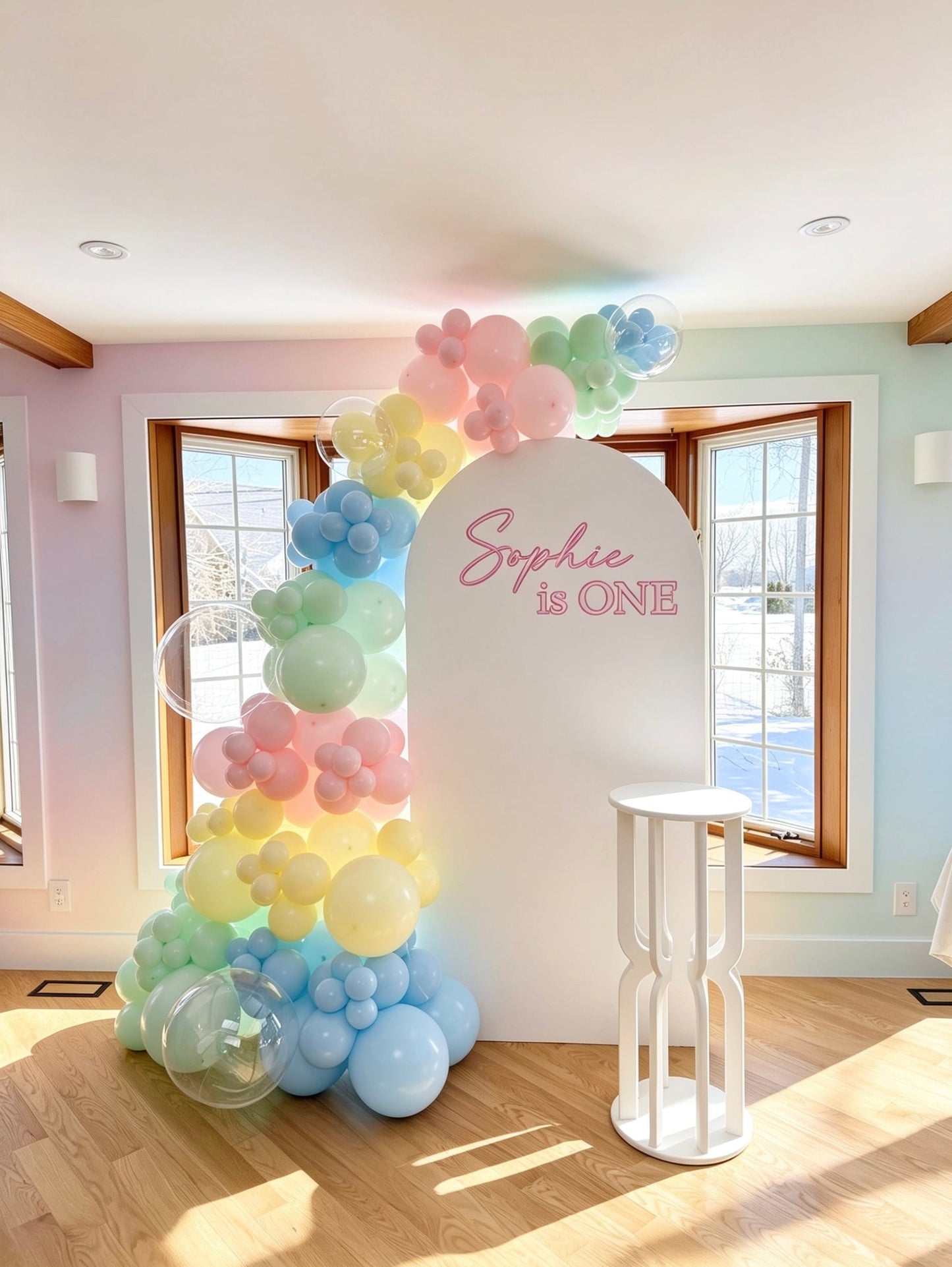 pastel rainbow first birthday setup with white full arch backdrop pastel balloons and cake table