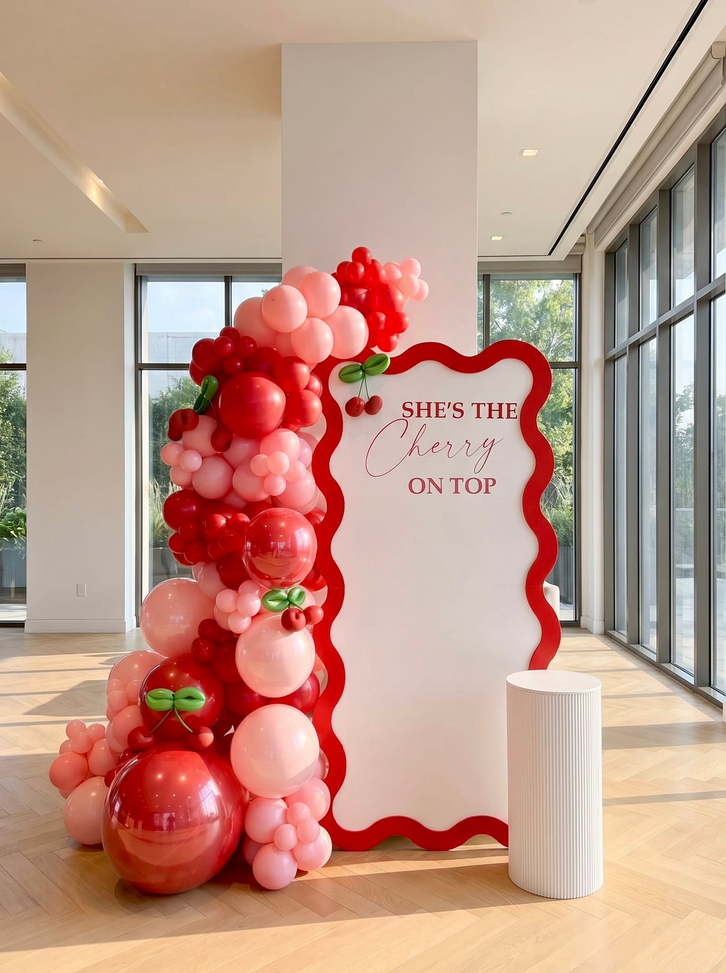 Cherry-themed birthday backdrop featuring a white wavy panel with red border, custom lettering, pink and red balloon garland, cherry accents, and a white ribbed pedestal stand.