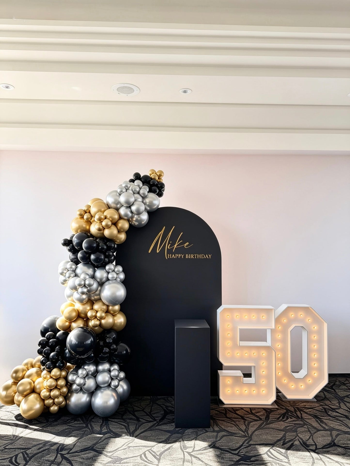 Black arch backdrop with black, gold and silver balloon garland and illuminated 50 marquee numbers for a 50th birthday celebration