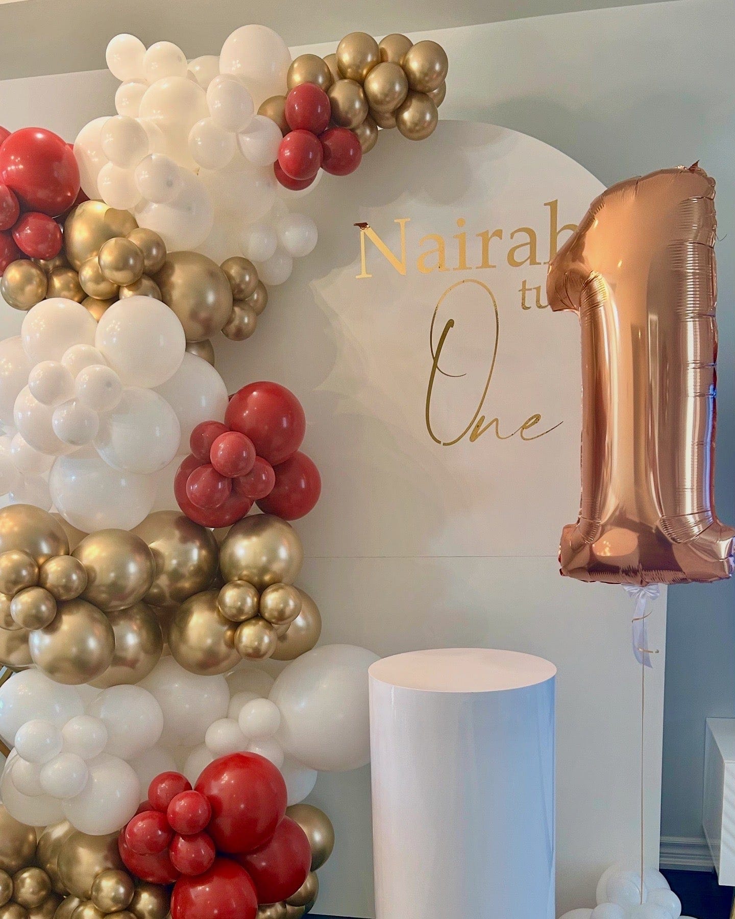 First birthday backdrop with white, gold, and soft red balloon garland, gold lettering, cake table, and floating number one balloon