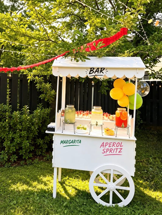 White outdoor cocktail bar cart with a scalloped canopy, decorative wheel, drink dispensers, and Margarita and Aperol Spritz lettering.