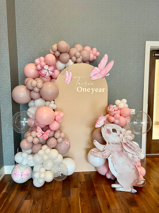 First birthday setup with beige backdrop, pink balloon garlands, butterfly accents and a bunny cut-out holding a butterfly.