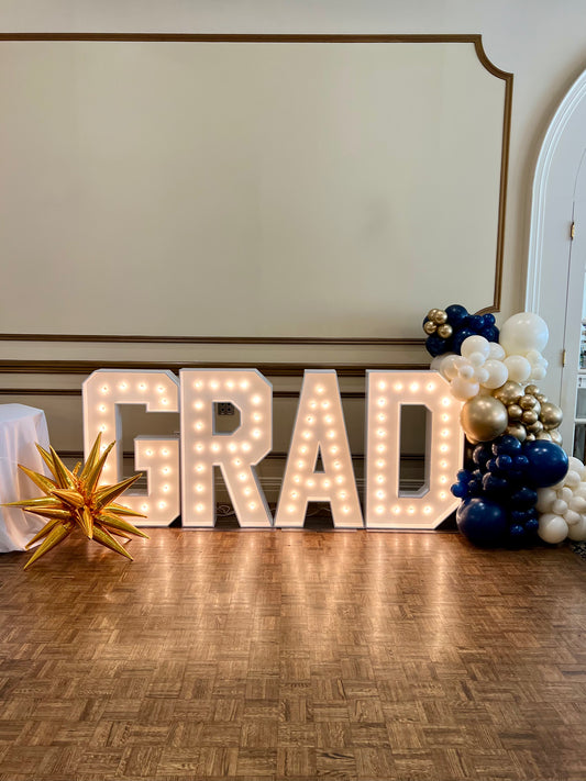Decorative 'GRAD' sign with lights, balloons, and a star-shaped object on a wooden floor.