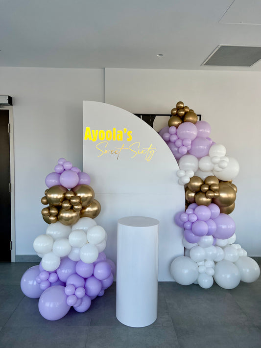 Birthday setup with half-circle backdrop, lavender and gold balloon garlands, custom inscription and cake table