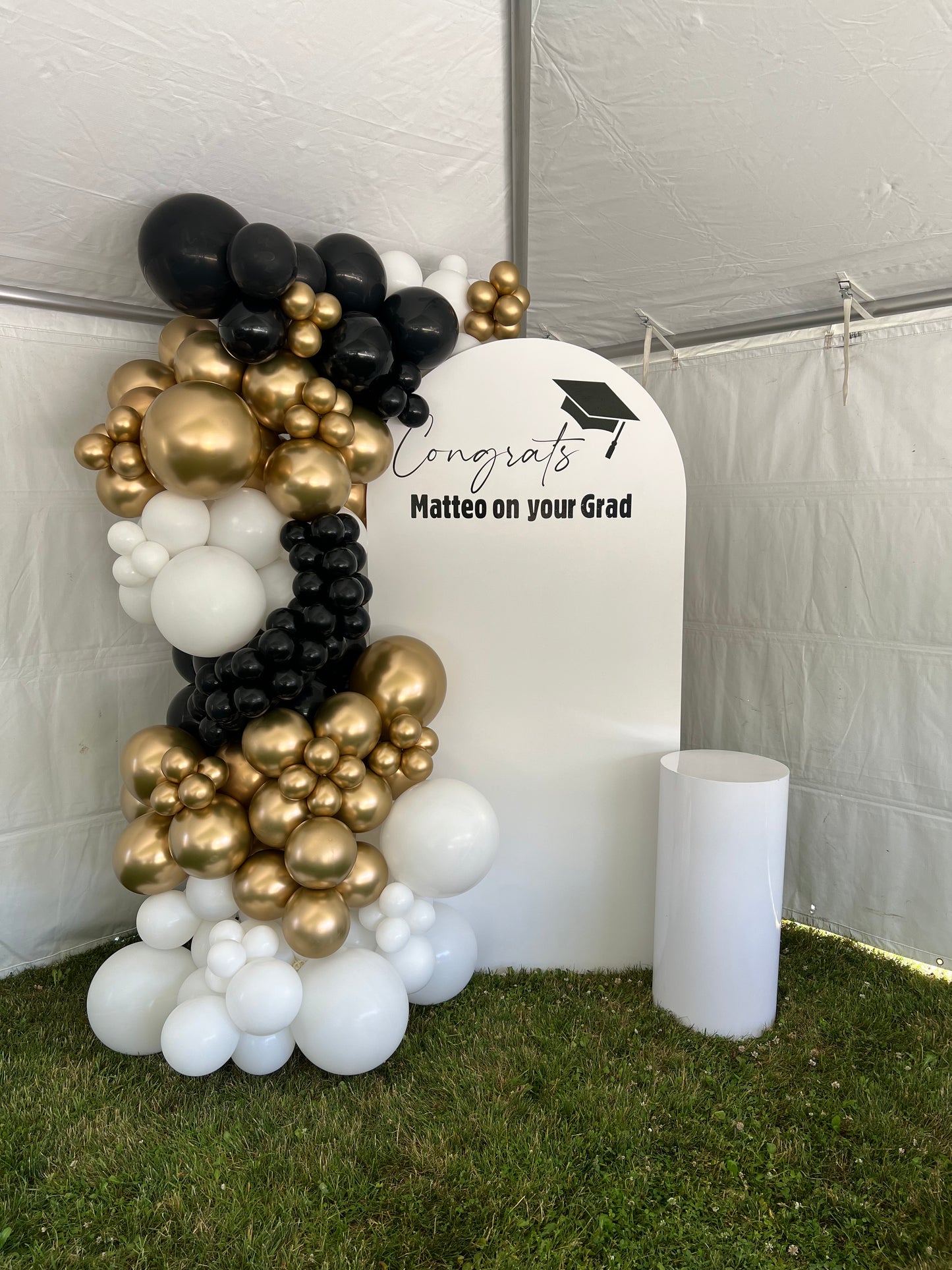 Graduation-themed balloon arch with 'Congrats! Matteo on your Grad' sign against a white wall.