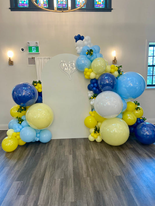 Balloon arch with blue, yellow, and white balloons in a room with a wooden floor and large windows.