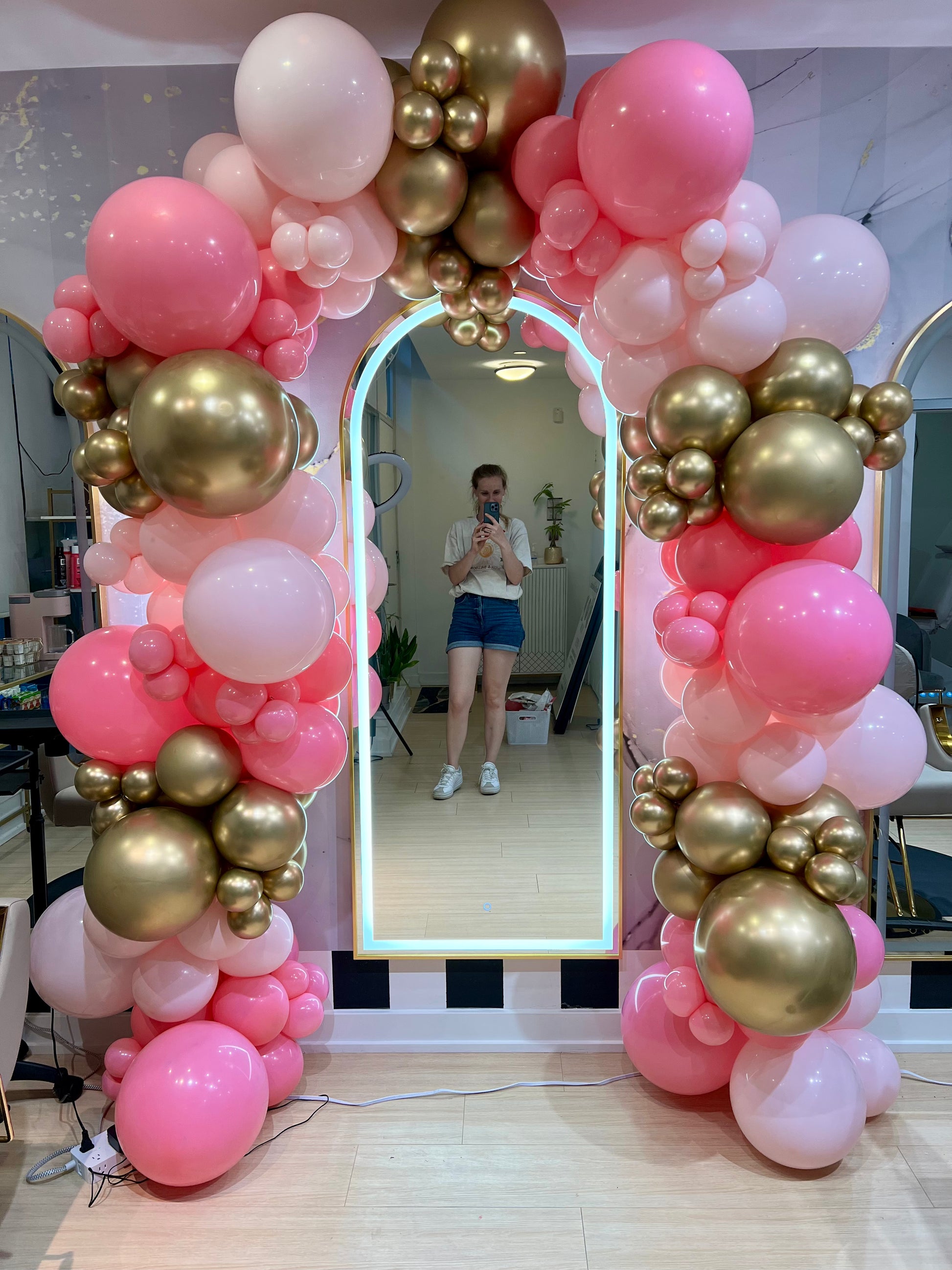 Person taking a mirror selfie in front of a decorative balloon arch with pink, gold, and white balloons.