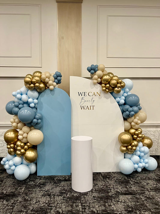We Can Bearly Wait baby shower backdrop with white and blue walls, beige gold & blue balloons, cake table, and custom sign
