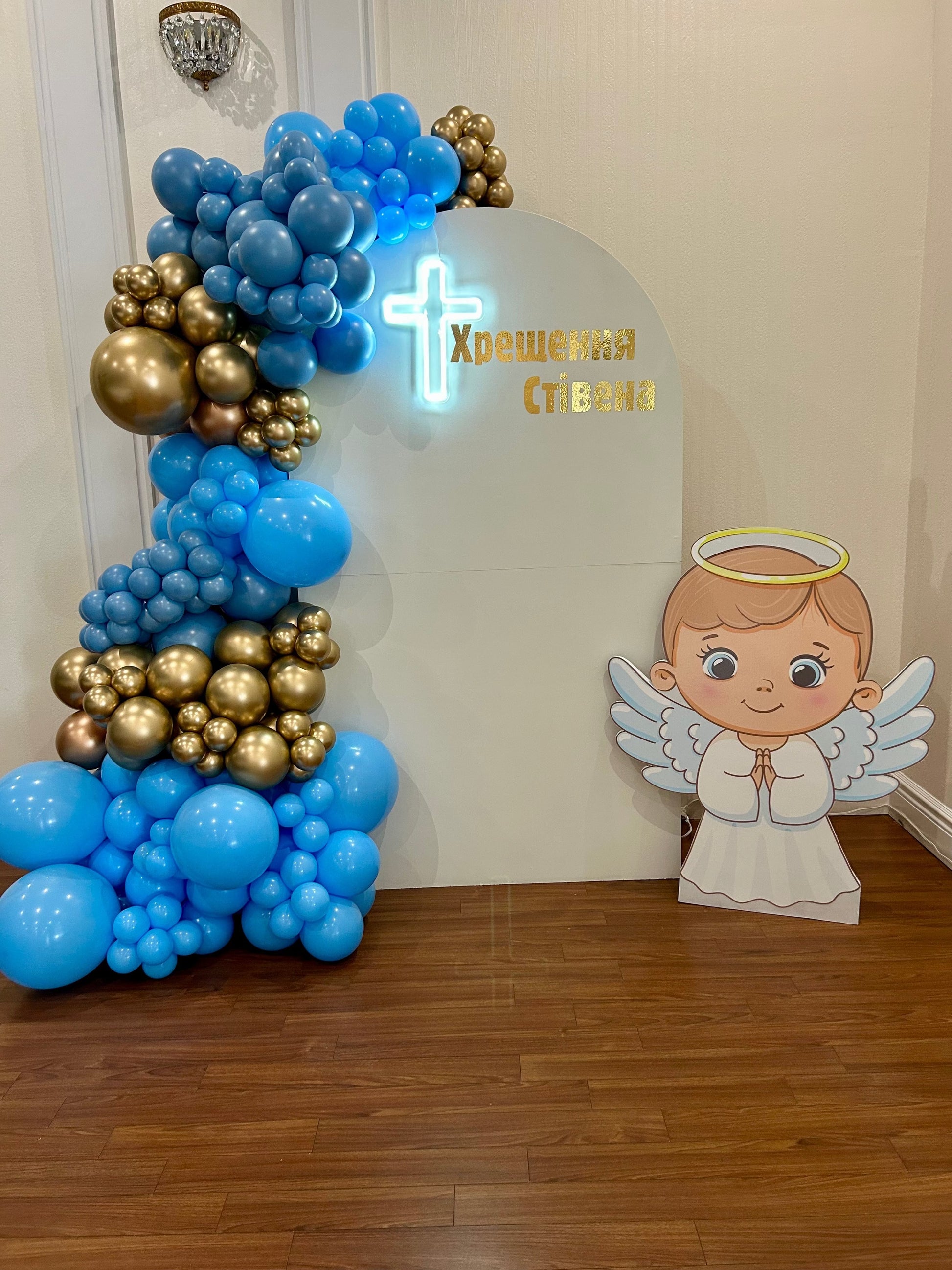 White baptism backdrop with blue and gold balloon garland, neon cross and angel cut-out
