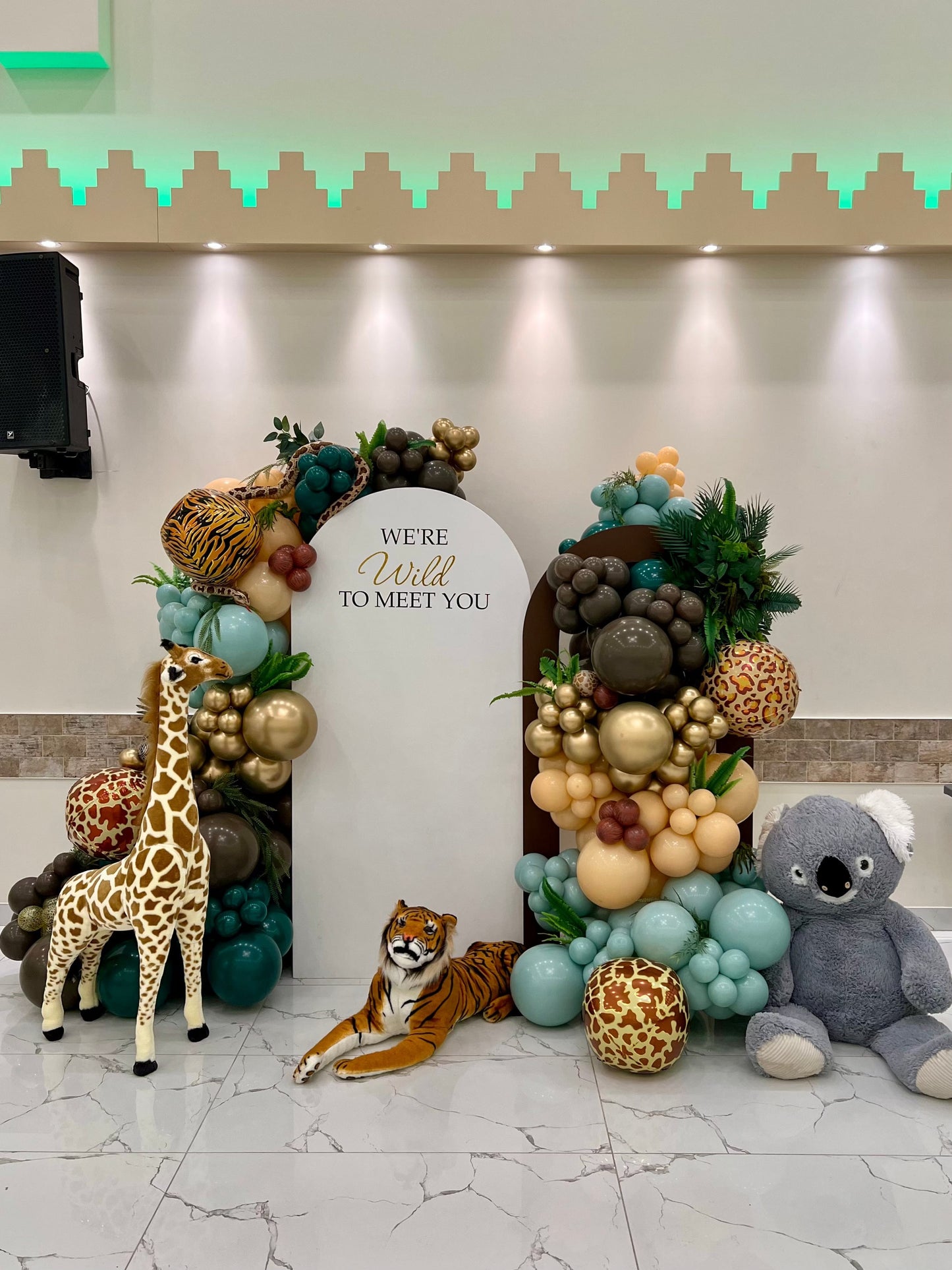 Safari baby shower backdrop with white and brown arches, balloon garlands, greenery, giraffe, tiger, snake, koala & custom inscription
