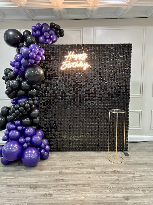 Birthday setup with black shimmer wall, purple and black balloons, gold cake table, and neon “Happy Birthday” sign