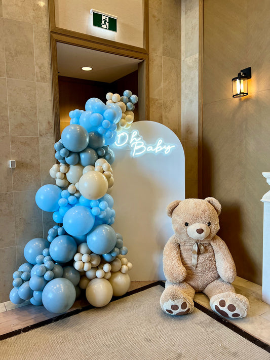 Blue baby shower setup with white full arch backdrop, Oh Baby neon sign, and giant teddy bear