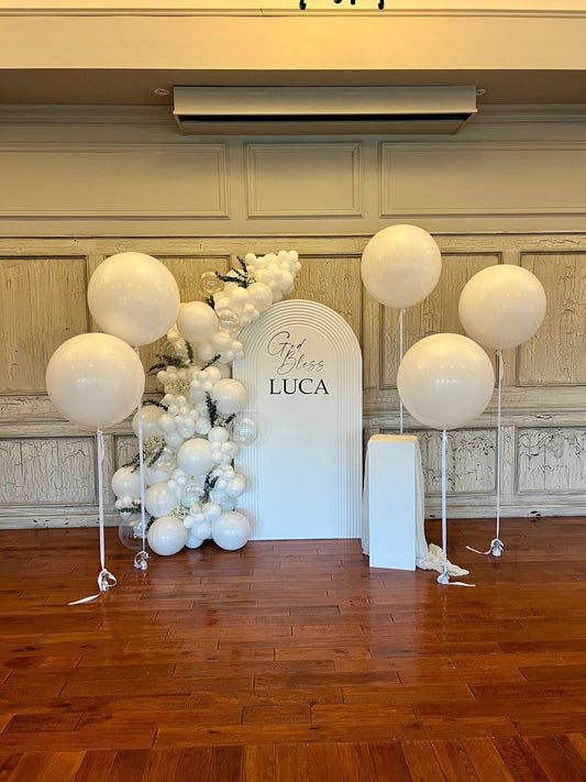 White baptism backdrop setup with floral balloon garland and large white balloons.