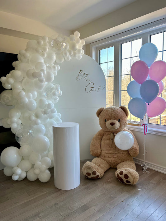 Gender reveal balloon backdrop with white garland, golden lettering, teddy bear, cake table, and pastel balloon bouquet

