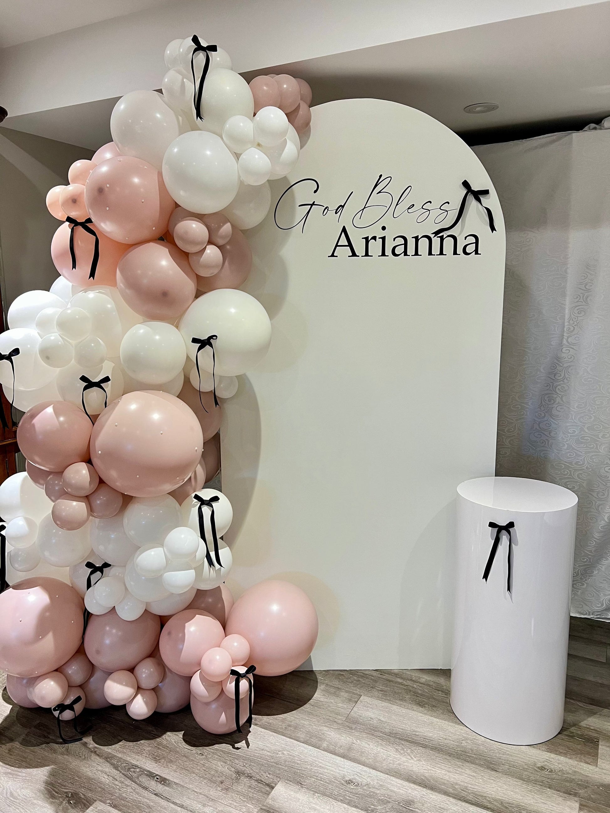 White oval baptism backdrop with “God Bless” name inscription, pink balloon garland, bows, pearls, and cake table