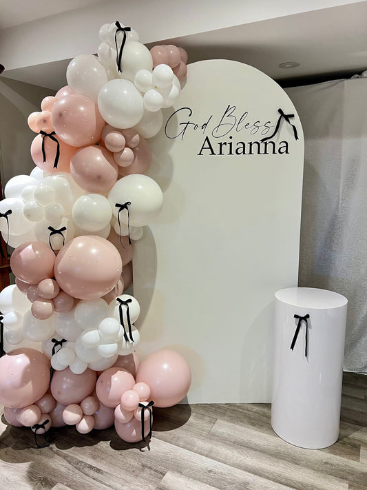 White oval baptism backdrop with “God Bless” name inscription, pink balloon garland, bows, pearls, and cake table