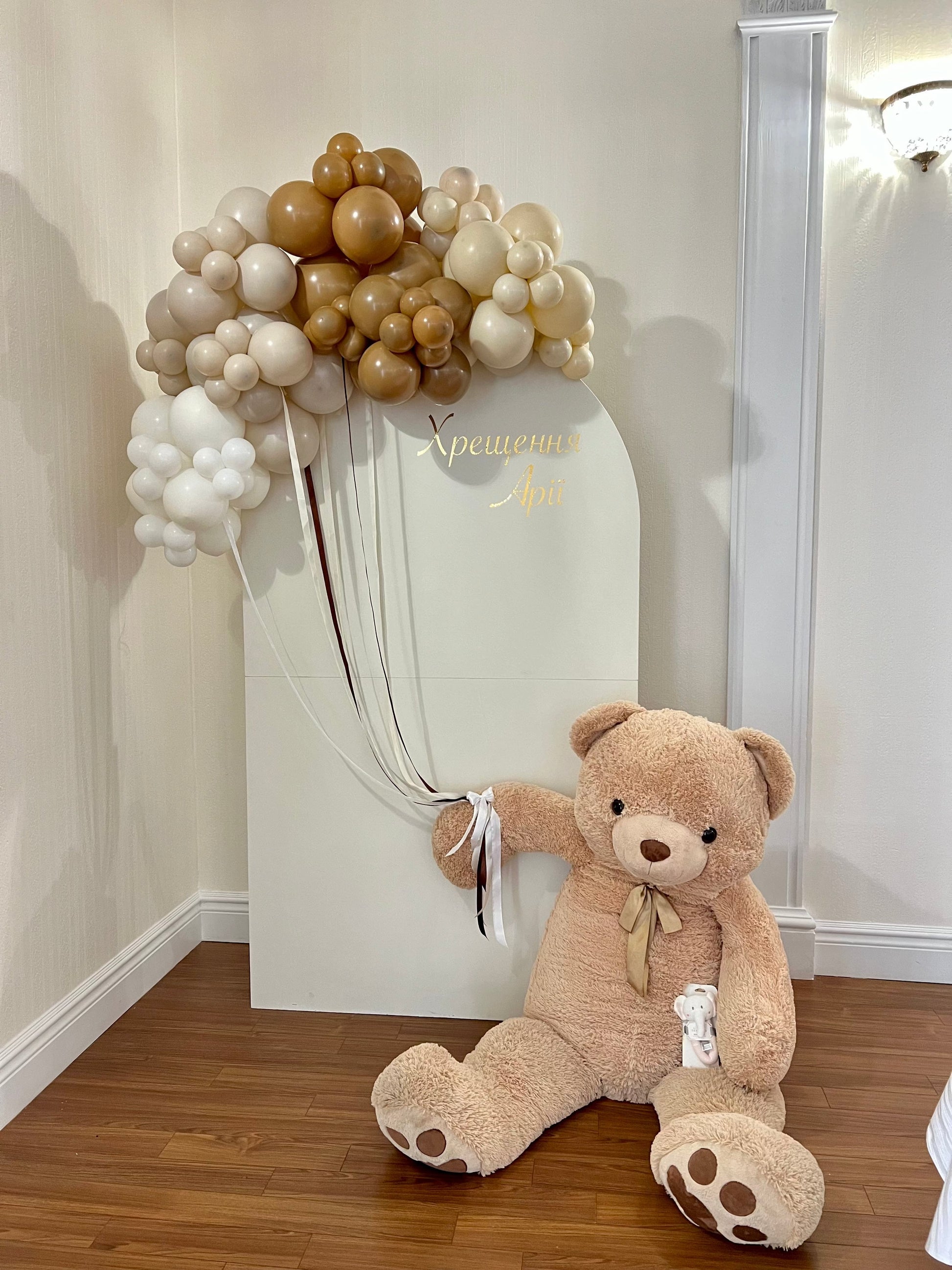 White oval baptism backdrop with teddy bear holding balloon strings, neutral balloon garland, custom inscription
