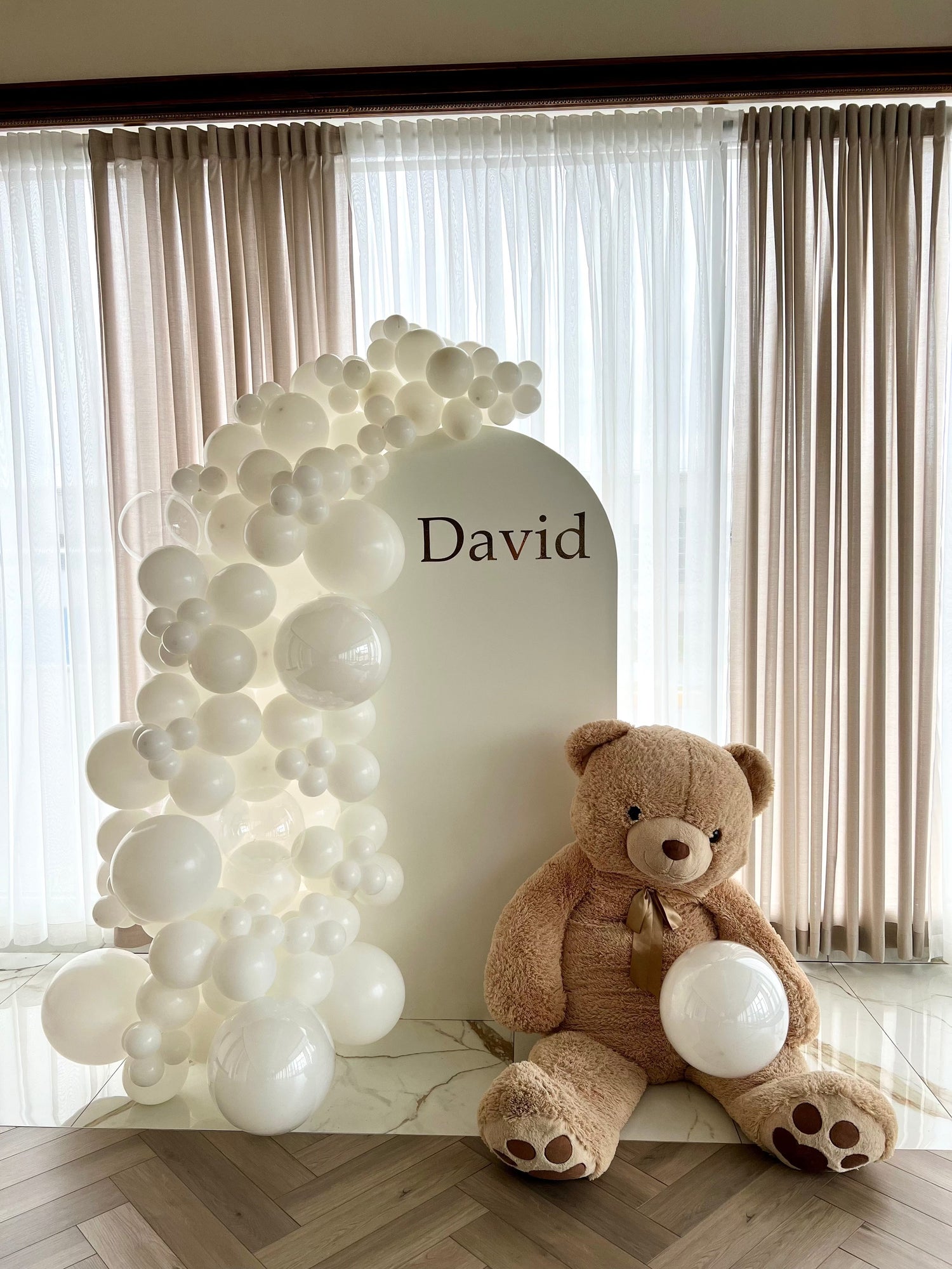 White oval backdrop with balloon garland, teddy bear prop, and custom child’s name inscription