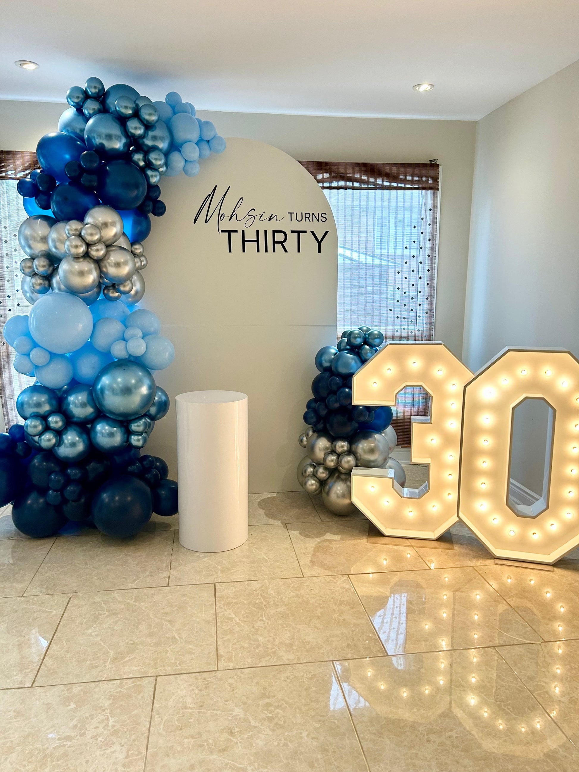 White oval birthday backdrop with custom inscription, blue & silver balloon garlands, marquee numbers 30, and white cake table
