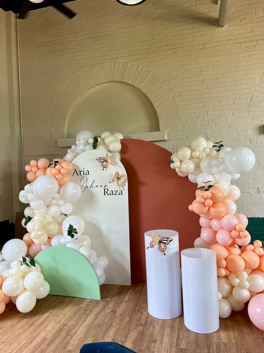 Neutral birthday setup with white, orange, and green backdrops, balloon garlands, butterflies, and custom vinyl sign