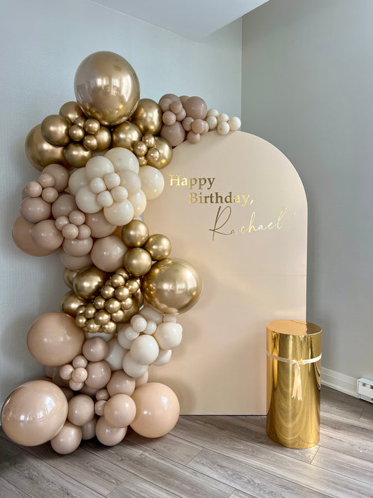 Warm neutral birthday setup with beige full arch backdrop, gold cake table and golden balloon garland.