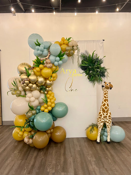 Jungle safari first birthday setup with white full arch backdrop, safari balloon garland, greenery, plush snake and giraffe prop.