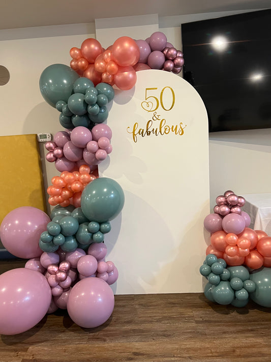 Elegant 50th birthday setup with white full arch backdrop, custom sign and balloon garlands in purple, green and orange tones.